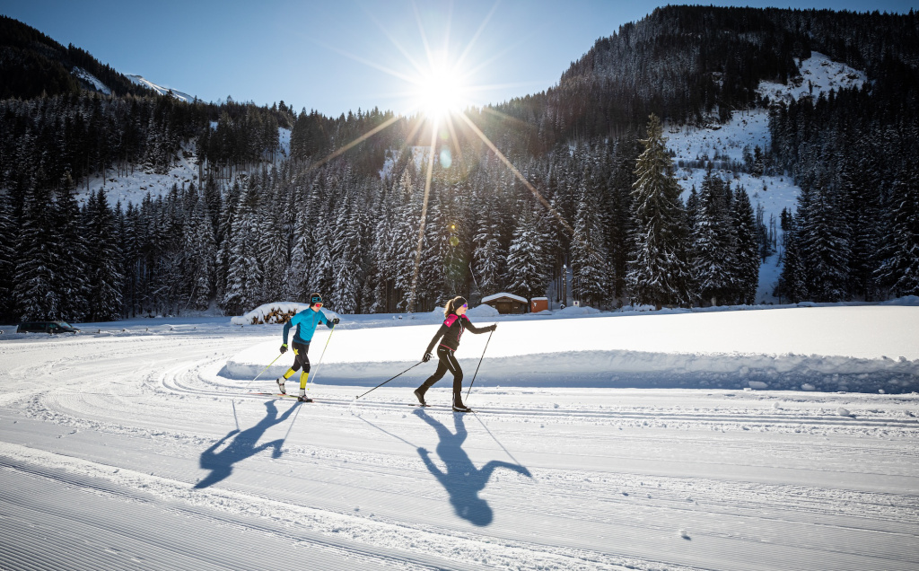 Two cross-country skiers on a groomed trail in front of a snow-covered coniferous forest in a sunny mountain valley